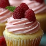 A close up of a lemon cupcake topped with a tall swirl of pink raspberry buttercream frosting and a fresh raspberry.
