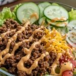 A colorful meal prep bowl containing ground beef, shredded lettuce, cheese, pickles, and a creamy orange dressing.