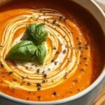 A bowl of creamy orange tomato soup topped with fresh herbs and cracked black pepper next to a spoon.
