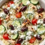 A colorful Greek orzo salad with diced chicken, feta cheese, and fresh vegetables in a large white bowl.