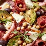 A large glass bowl filled with Italian Antipasto Salad including salami, mozzarella pearls, and artichokes.