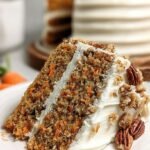 A slice of healthy carrot cake with thick white yogurt frosting and chopped walnuts on a white plate.