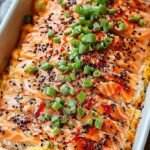 A golden-brown salmon sushi bake in a glass dish topped with green onions and sesame seeds