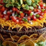 A colorful seven-layer taco salad in a glass dish with layers of beans, cream, and fresh vegetables.