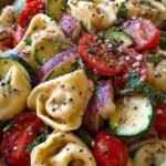A colorful tortellini pasta salad with cherry tomatoes, cucumbers, and feta cheese in a glass bowl