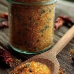 A glass jar filled with a vibrant red and black Homemade Cajun Seasoning blend on a wooden kitchen counter.