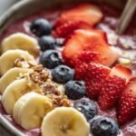 A thick pink strawberry banana smoothie bowl topped with fresh fruit, granola, and chia seeds in a white bowl.