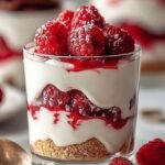 A glass jar layered with creamy yogurt, fresh red raspberries, and golden granola.