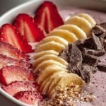 A thick pink strawberry smoothie bowl topped with fresh berries, granola, and seeds in a white bowl.