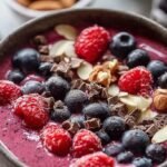 A thick purple berry smoothie bowl topped with banana slices, chia seeds, and granola.