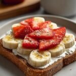 A slice of whole grain toast topped with creamy cottage cheese, sliced strawberries, bananas, honey, and cinnamon.