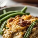A white baking dish filled with creamy green beans topped with golden crushed pork rinds and parmesan cheese.