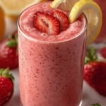 A bright pink strawberry lemonade smoothie in a chilled glass garnished with a lemon slice.
