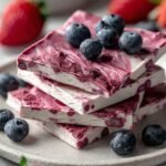 A tray of frozen yogurt bark topped with fresh strawberries, blueberries, granola, and hemp seeds broken into shards.