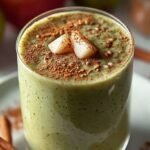 A tall glass of creamy cinnamon pear smoothie topped with a dash of ground cinnamon and a fresh pear slice on a wooden table.