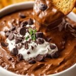 A bowl of creamy chocolate pumpkin dip served with apple slices and pretzels on a wooden board.