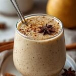 A glass of creamy spiced pear smoothie topped with a dash of cinnamon next to fresh pears.