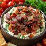 A layered creamy BLT dip in a white dish topped with bacon, tomatoes, and lettuce.