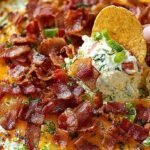 A layered creamy BLT dip topped with fresh lettuce, tomatoes, and crispy bacon bits in a white serving dish.