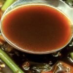 A glossy brown garlic stir fry sauce in a small saucepan with fresh ginger and garlic nearby.