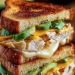 A golden-brown sourdough sandwich with melted cheese, sliced avocado, and chipotle chicken on a wooden board.